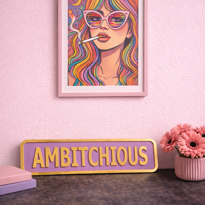 Framed artwork of a woman with colorful hair and sunglasses on a pink wall, with a 'AMBITIONOUS' sign below.