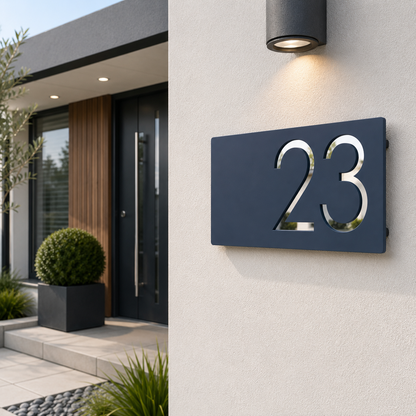 A modern navy blue house number sign displaying '23' with reflective silver numbers, mounted on a light gray stucco wall under a black outdoor wall light.
