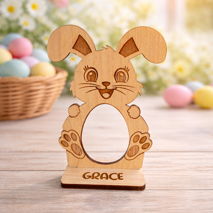 Wooden bunny-shaped photo frame with 'GRACE' engraving on a wooden surface with Easter eggs and flowers in the background.