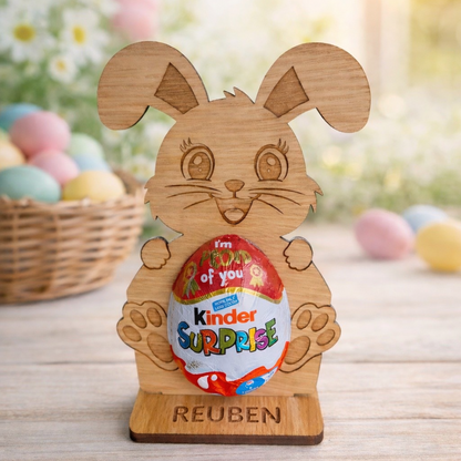 Wooden rabbit holder with a Kinder Surprise egg on a wooden surface with Easter eggs in the background.