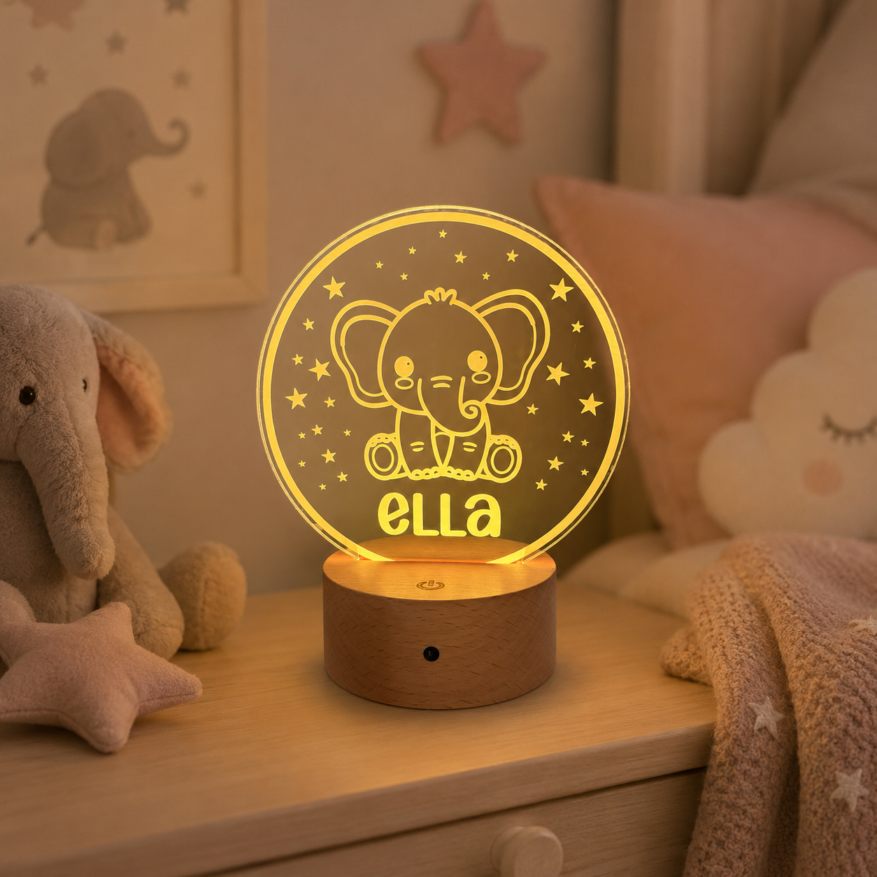 A personalized 3D LED night light shaped like a sleeping elephant, glowing with a warm golden-yellow light on a wooden base. The name 'ella' is custom engraved on the front.