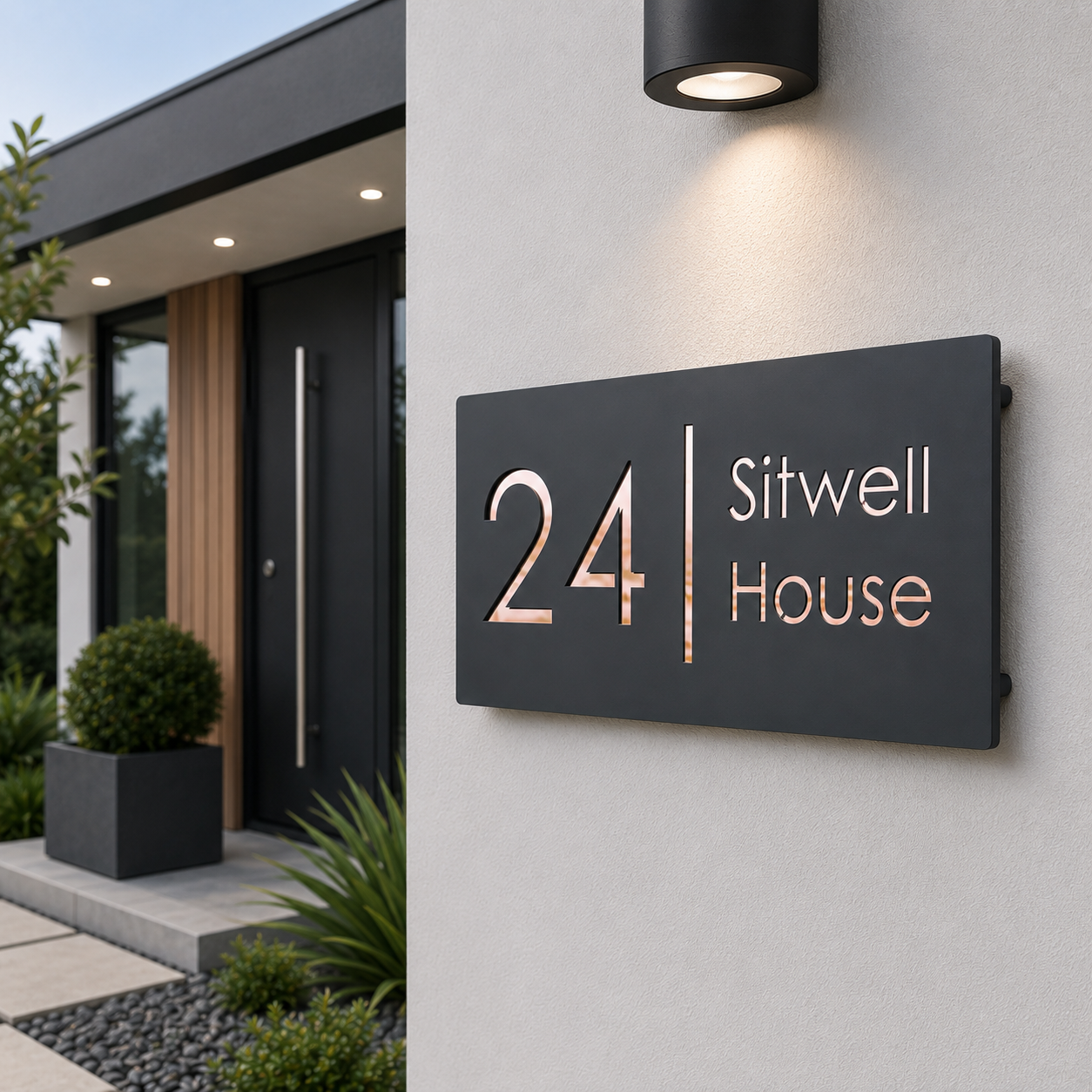 A modern black metal house number sign reading '24 Sitwell House' mounted on a light gray exterior wall under a black wall light. The sign features rose gold numerals and a vertical divider, with white lettering for 'Sitwell House'.