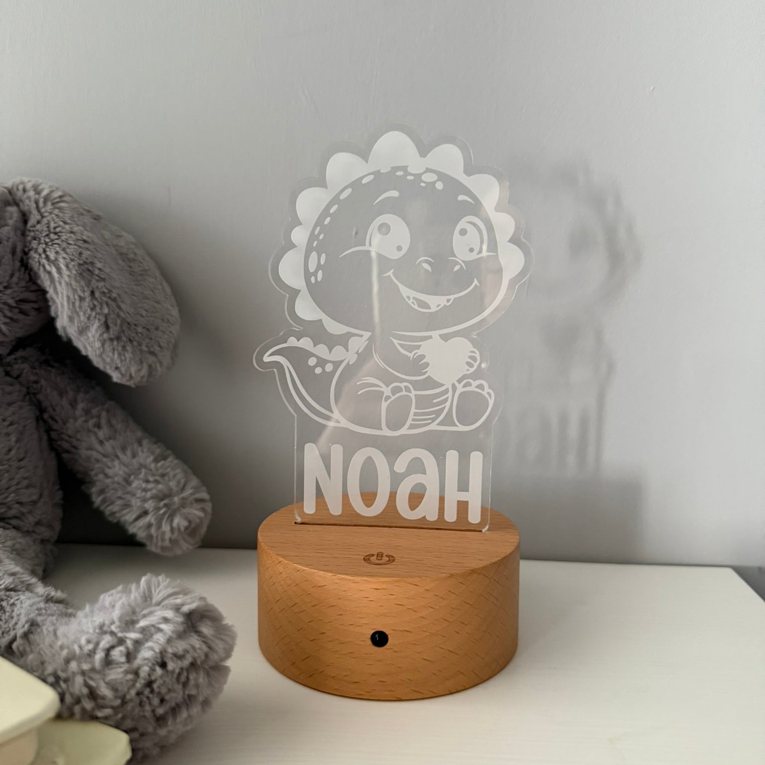 A personalized acrylic LED night light featuring a cartoon dinosaur and the name 'NOAH' on a round wooden base, styled on a white surface next to a gray plush toy.