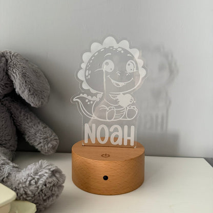 A personalized acrylic LED night light featuring a cartoon dinosaur and the name 'NOAH' on a round wooden base, styled on a white surface next to a gray plush toy.