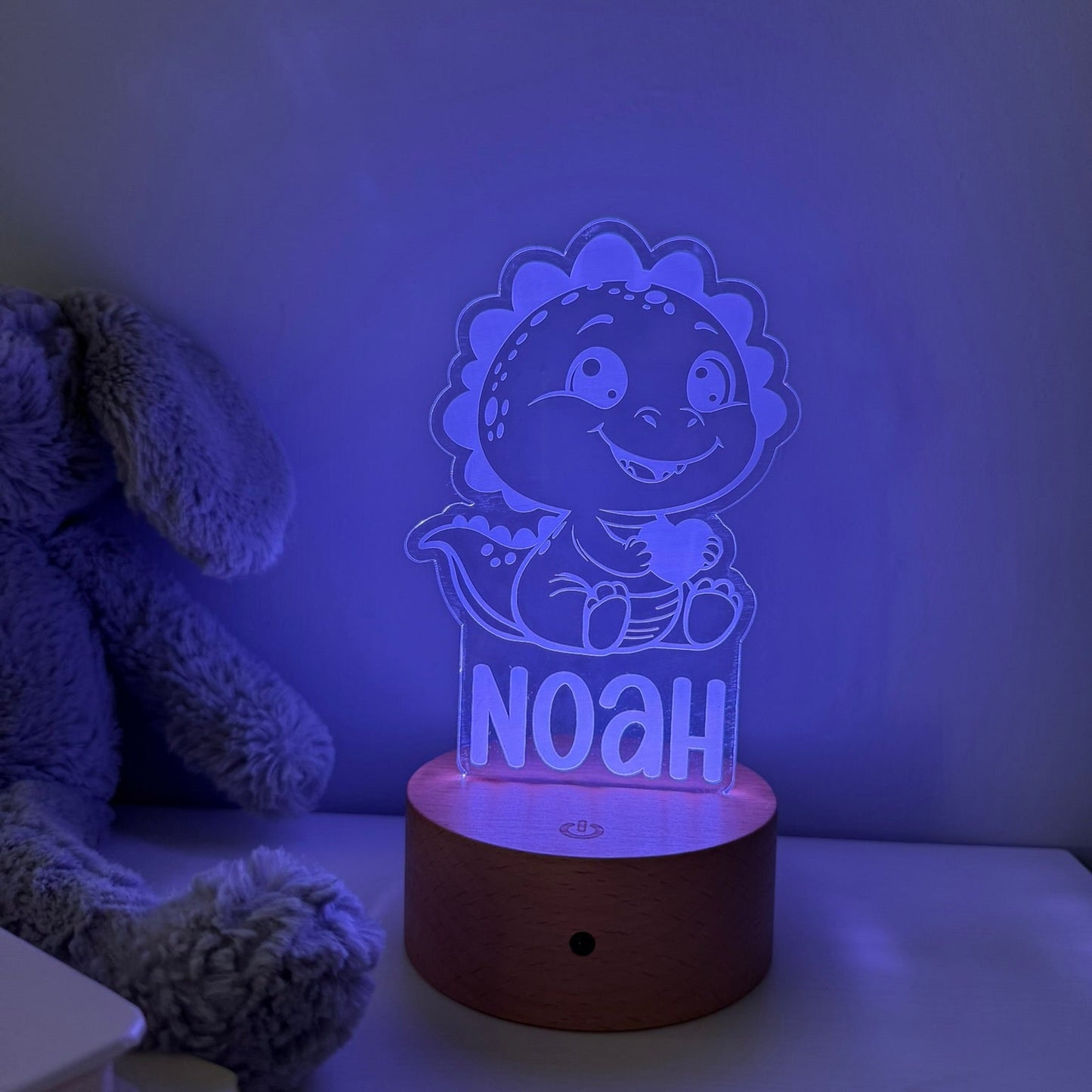 A personalized LED night light featuring a cute cartoon dinosaur holding a cup with the name 'NOAH' on it, sitting on a round wooden base. The scene is illuminated with a blue-purple glow, styled next to a plush toy on a shelf.