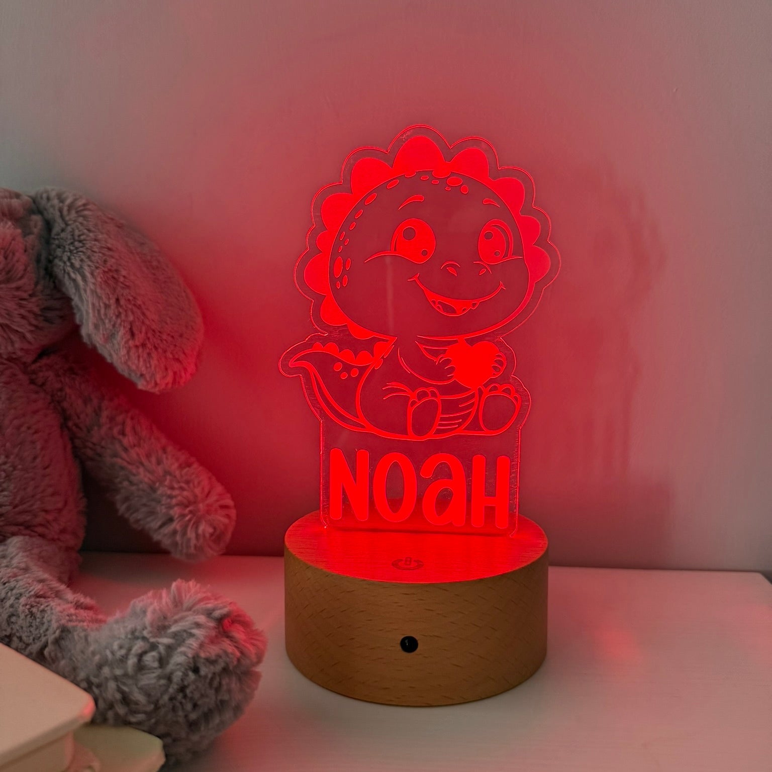 A personalized LED night light shaped like a cartoon dinosaur with the name 'NOAH' illuminated on the front, sitting on a round wooden base on a tabletop next to a plush toy.