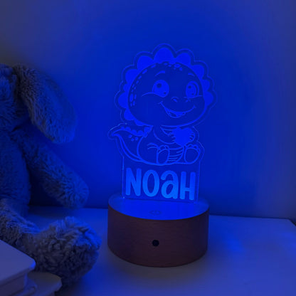A personalized 3D acrylic night light featuring a cartoon dinosaur and the name 'NOAH' on a wooden base, glowing with a blue light in a bedroom setting next to a plush toy.