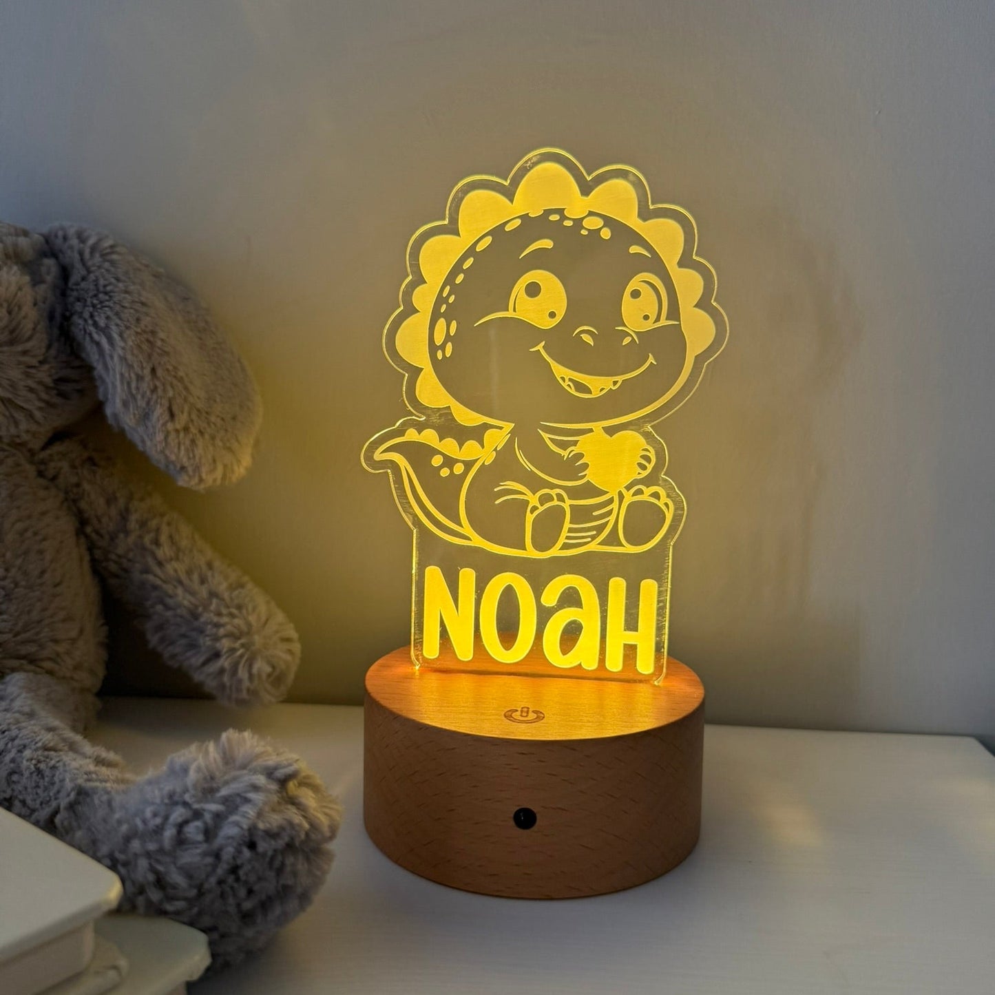 A personalized LED night light shaped like a cartoon dinosaur sitting on a wooden base, glowing with warm yellow light. The name 'NOAH' is printed on the front.