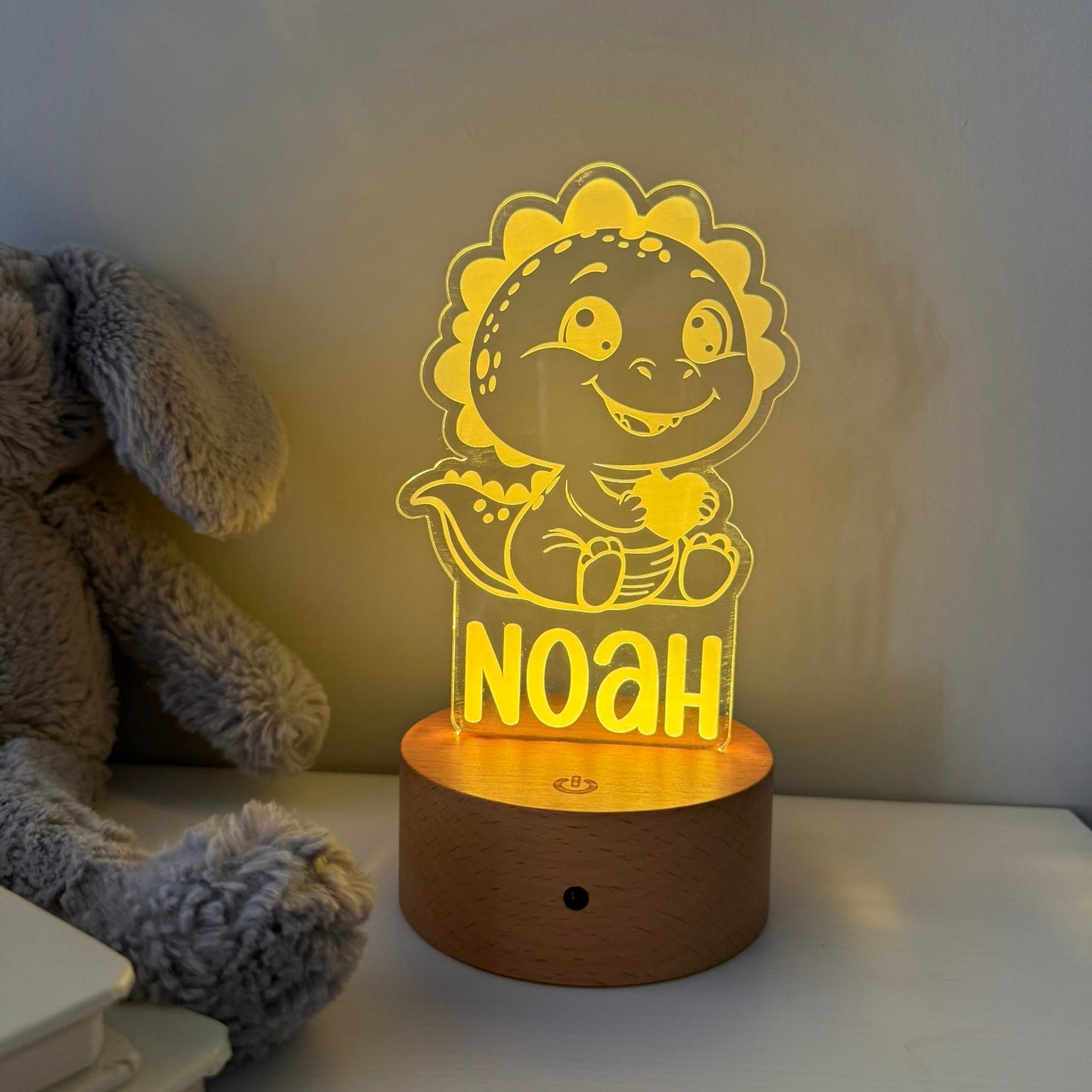 A personalized LED night light shaped like a cartoon dinosaur sitting on a wooden base, glowing with warm yellow light. The name 'NOAH' is printed on the front.
