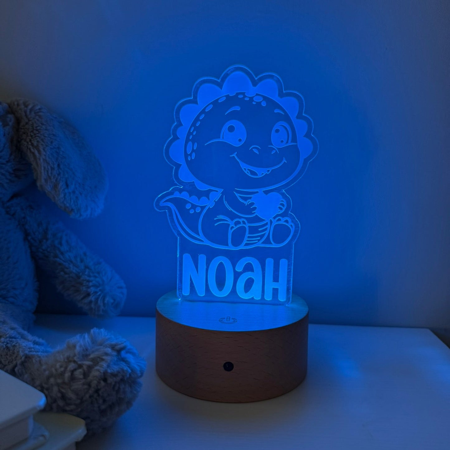 A personalized LED night light featuring a cartoon dinosaur character and the name 'NOAH' on a glowing acrylic panel, set on a wooden base on a tabletop next to a plush toy.