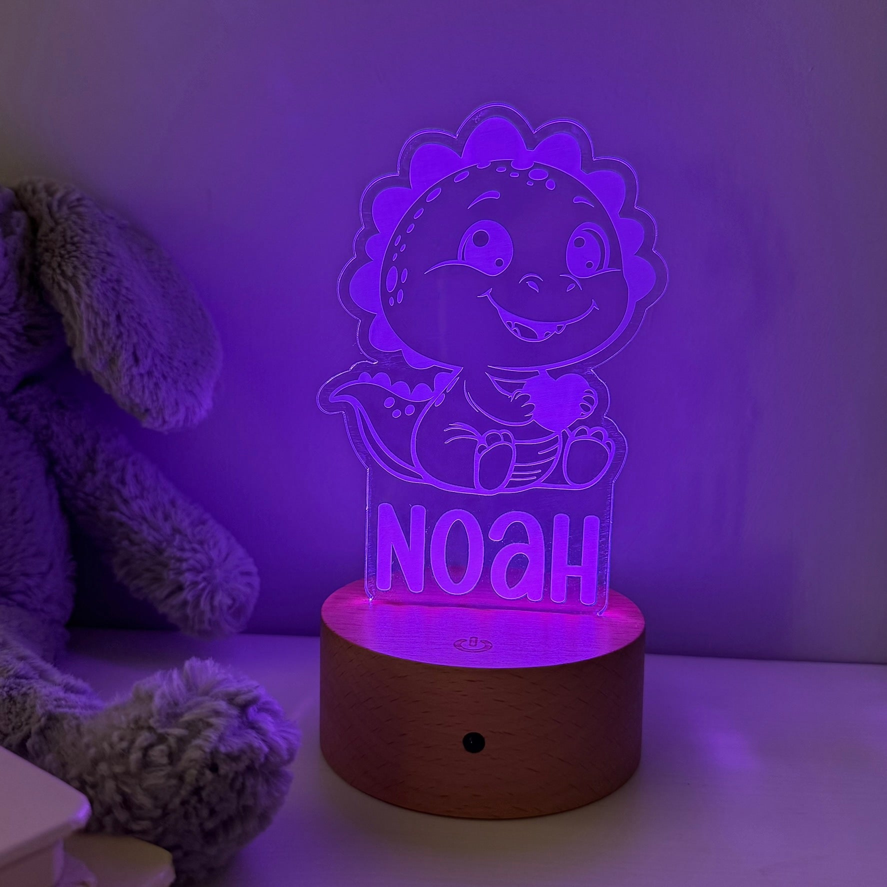 A personalized 3D acrylic LED night light shaped like a smiling cartoon dinosaur holding a heart, with the name 'NOAH' illuminated on the front, resting on a round wooden base. The lamp is glowing with a purple light in a bedroom setting next to a plush toy.