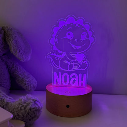 A personalized 3D acrylic LED night light shaped like a smiling cartoon dinosaur holding a heart, with the name 'NOAH' illuminated on the front, resting on a round wooden base. The lamp is glowing with a purple light in a bedroom setting next to a plush toy.