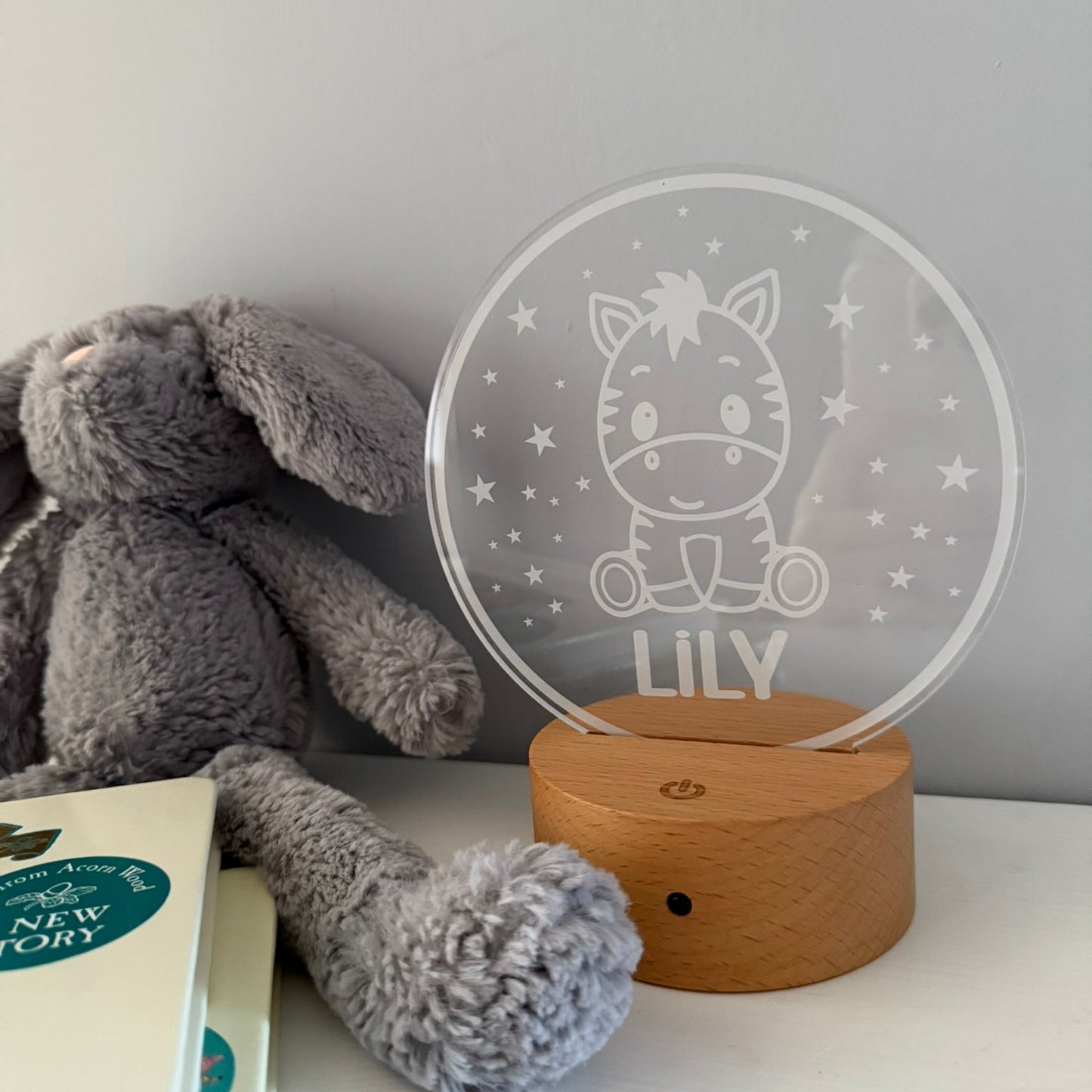 A personalized acrylic night light with a zebra design and the name 'LILY' on a wooden base, styled next to a gray plush bunny on a white surface.