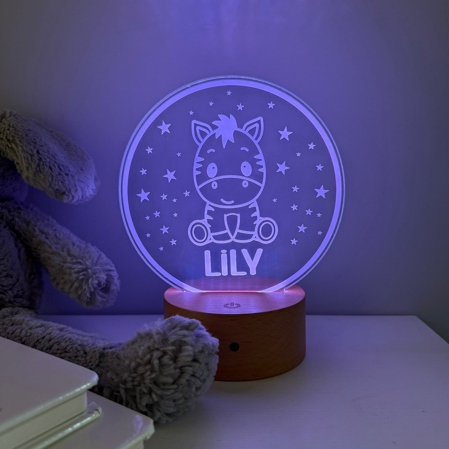 A personalized 3D optical illusion night light shaped like a circular snow globe, featuring a cartoon zebra on a tricycle and the name 'LILY' illuminated in purple. It sits on a wooden base on a bedside table next to a plush toy and books.