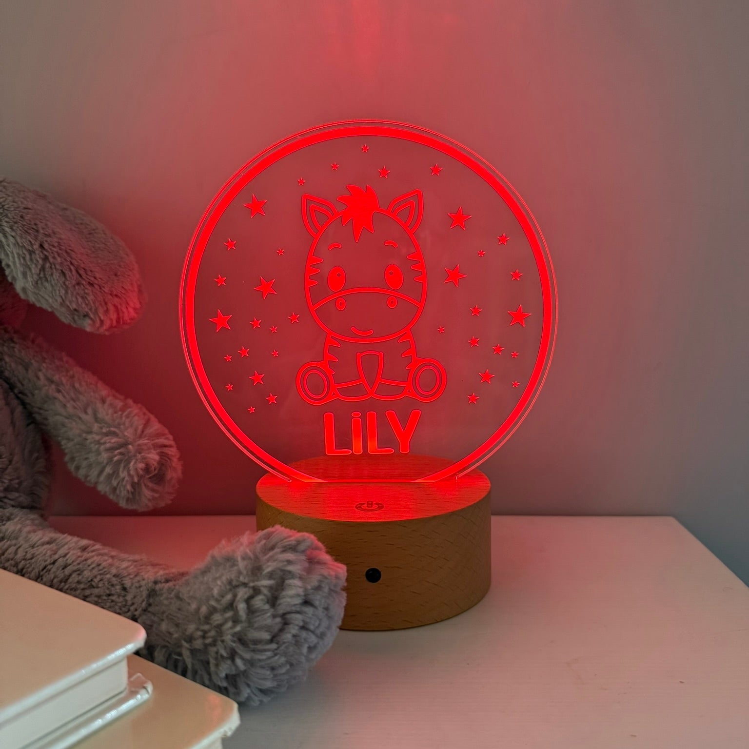 A personalized acrylic 3D LED night light shaped like a cat sitting in a wheel, glowing red with the name 'LILY' on it, resting on a wooden base on a white table next to a gray plush toy and books.