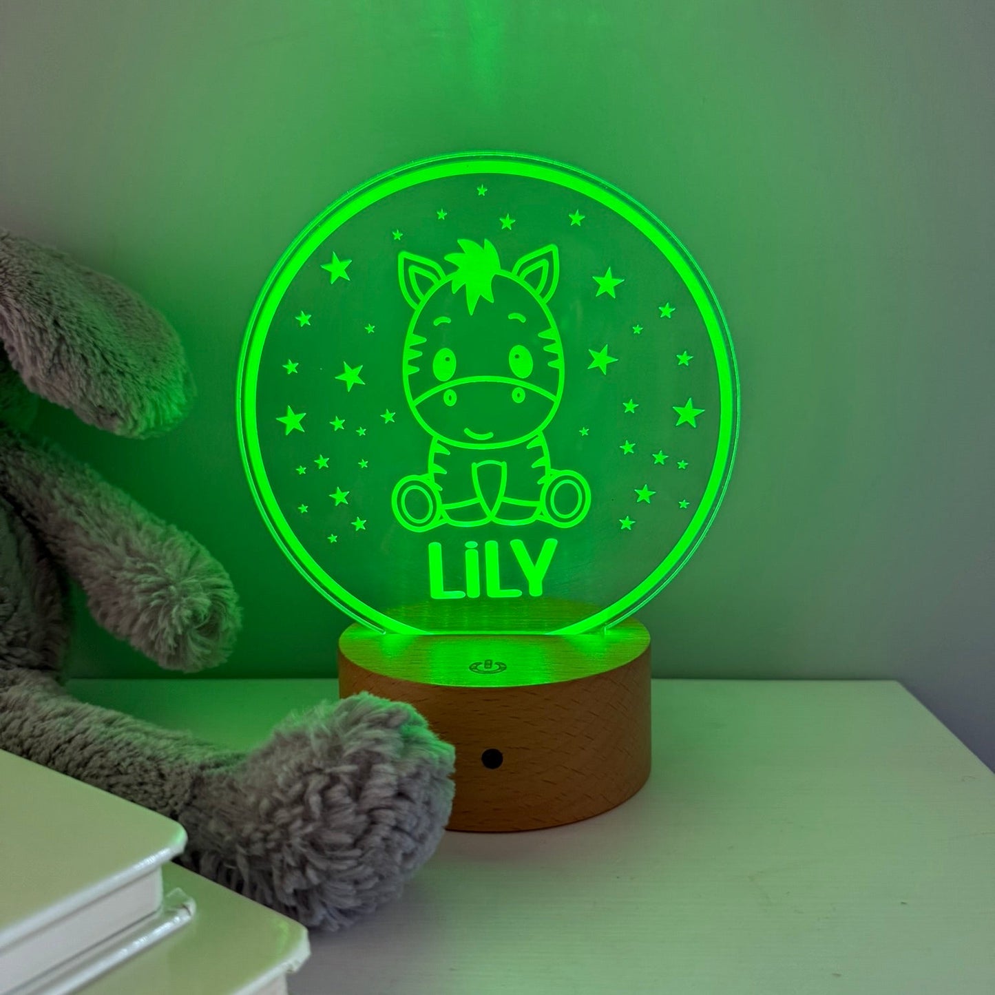 A personalized 3D optical illusion night light shaped like a round glowing green crystal ball with a cartoon horse and the name 'LILY' on a wooden base, shown on a tabletop next to a plush toy.