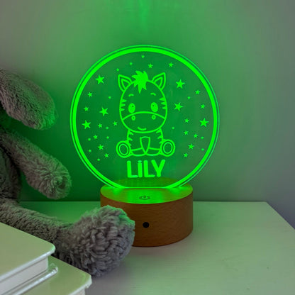 A personalized 3D optical illusion night light shaped like a round glowing green crystal ball with a cartoon horse and the name 'LILY' on a wooden base, shown on a tabletop next to a plush toy.