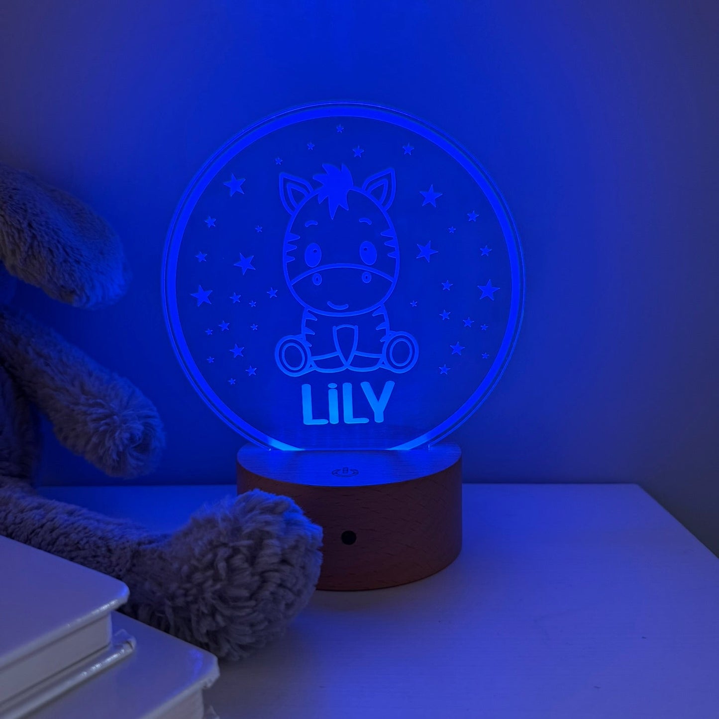 A personalized 3D optical illusion night light shaped like a round zebra car, glowing with a blue night-sky design and the name 'LILY' on a wooden base, styled on a tabletop next to a teddy bear and books.