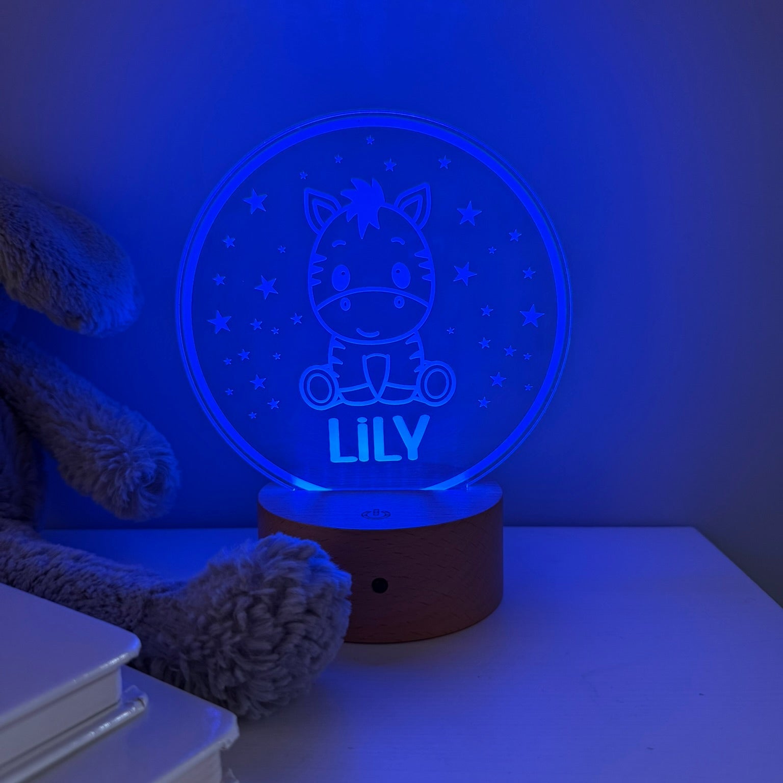 A personalized 3D optical illusion night light shaped like a round zebra car, glowing with a blue night-sky design and the name 'LILY' on a wooden base, styled on a tabletop next to a teddy bear and books.