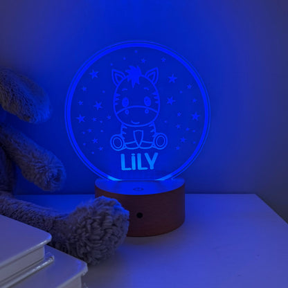 A personalized 3D optical illusion night light shaped like a round zebra car, glowing with a blue night-sky design and the name 'LILY' on a wooden base, styled on a tabletop next to a teddy bear and books.