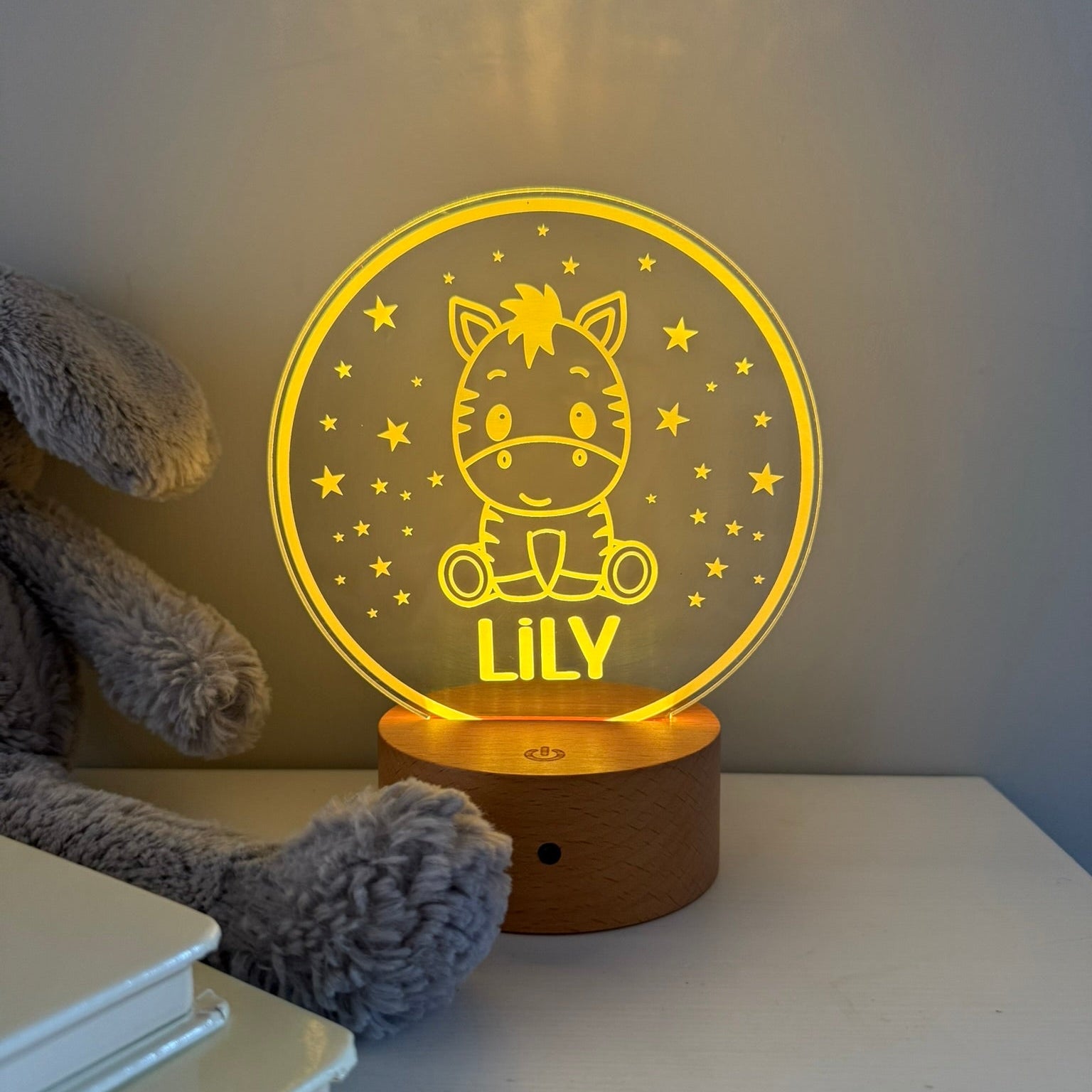 A personalized 3D acrylic night light shaped like a round moon with a cartoon cat and the name 'LILY' in glowing yellow, set on a wooden base on a white table next to a gray plush toy.
