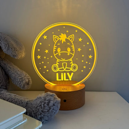 A personalized 3D acrylic night light shaped like a round moon with a cartoon cat and the name 'LILY' in glowing yellow, set on a wooden base on a white table next to a gray plush toy.