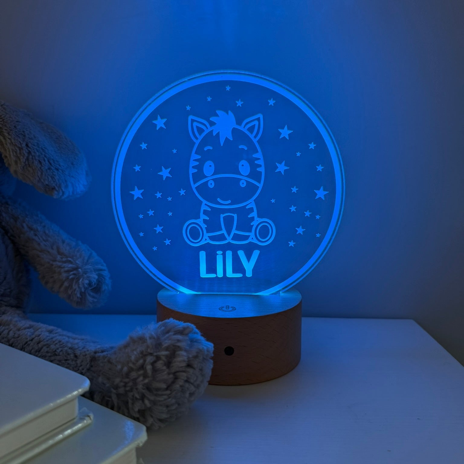 A personalized 3D optical illusion night light shaped like a round window, glowing with a blue light. The design features a cartoon zebra sitting in a starry sky with the name 'LILY' written below it.