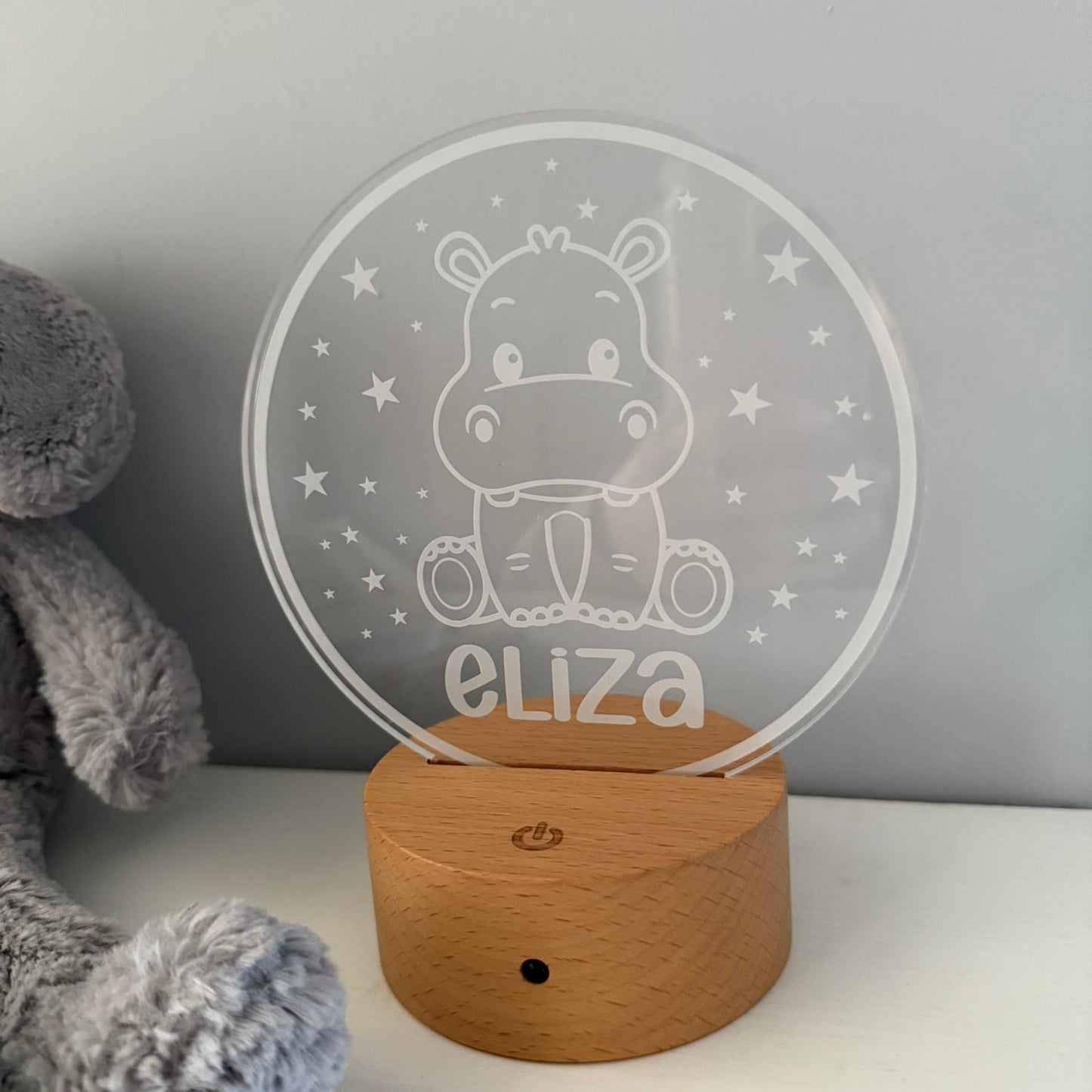 A personalized acrylic night light with a cartoon hippo and the name 'eliza' on a round wooden base, shown on a white surface next to a gray plush toy.