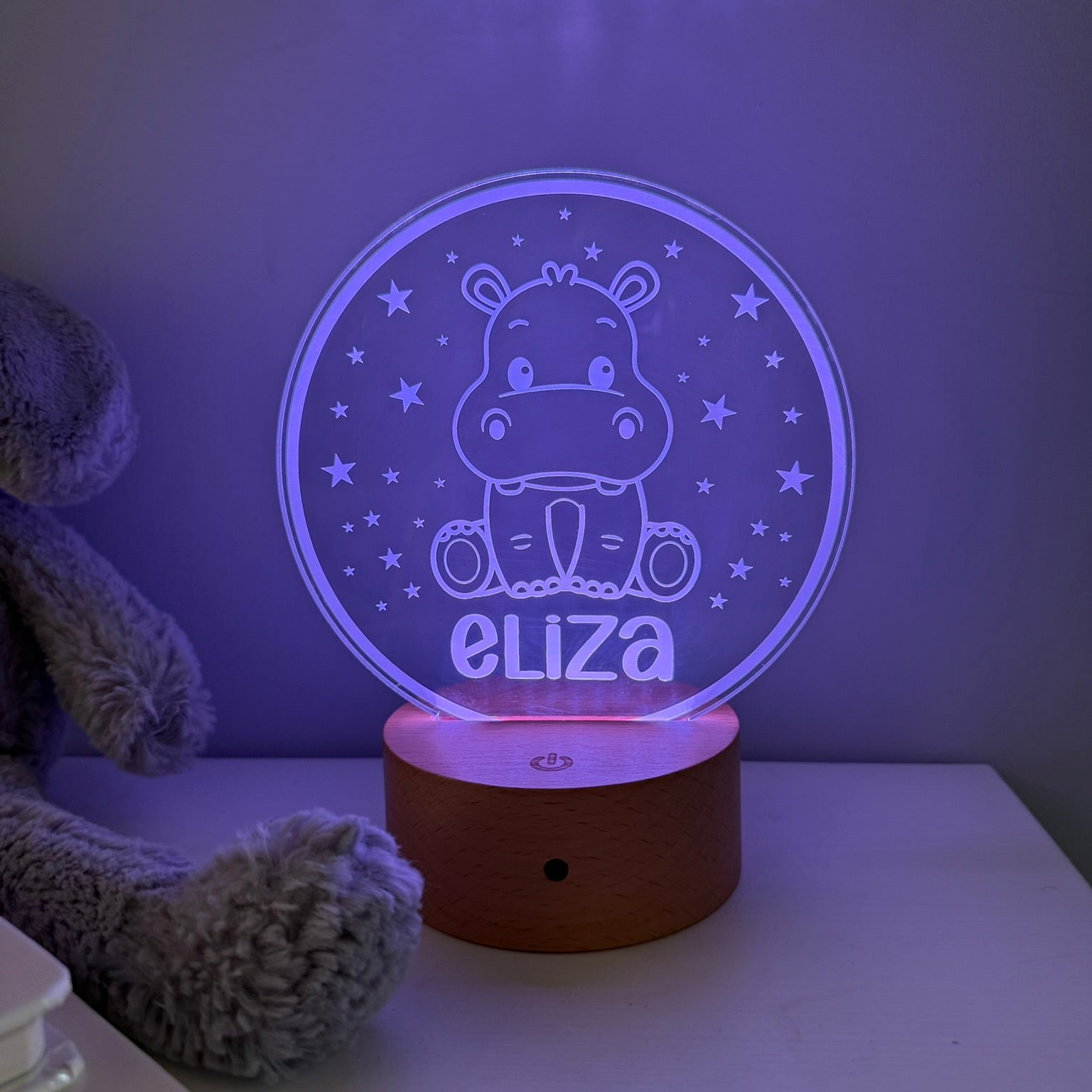 A personalized 3D acrylic night light featuring a cartoon hippo and the name 'eliza' in glowing purple light, resting on a wooden base on a white table next to a plush toy.