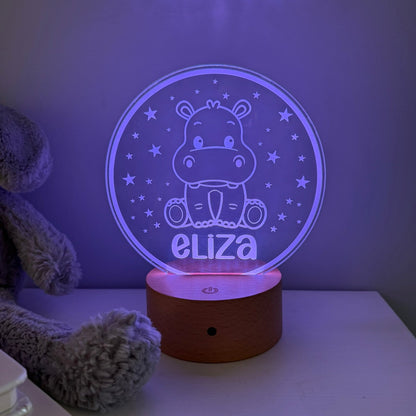 A personalized 3D acrylic night light featuring a cartoon hippo and the name 'eliza' in glowing purple light, resting on a wooden base on a white table next to a plush toy.