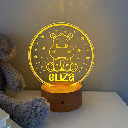 A personalized 3D acrylic night light shaped like a hippo sitting in stars, glowing with a warm yellow light. The name 'eliza' is custom engraved on the front, and the lamp sits on a wooden base on a white tabletop next to a gray plush toy.