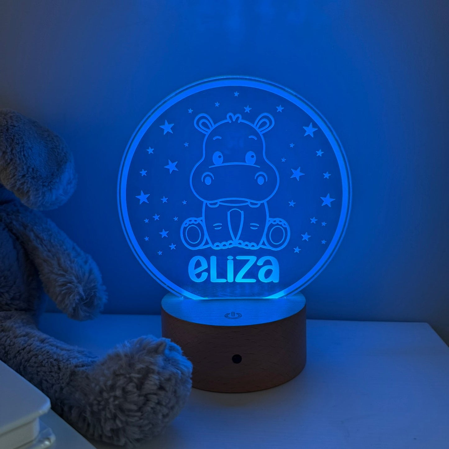 A personalized 3D acrylic night light shaped like a hippo sitting in a circle with stars, glowing blue with the name 'eliza' at the bottom, resting on a wooden base on a bedside table next to a plush toy.