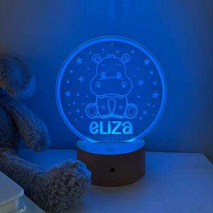 A personalized 3D acrylic night light shaped like a hippo sitting in a circle with stars, glowing blue with the name 'eliza' at the bottom, resting on a wooden base on a bedside table next to a plush toy.