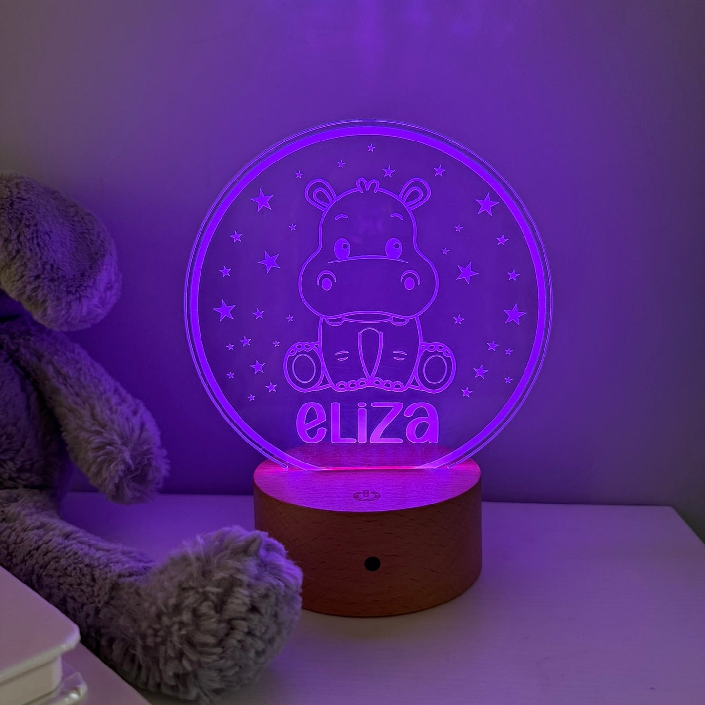 A personalized 3D optical illusion night light shaped like a hippo sitting in a circle, glowing with purple light and displaying the name 'eliza'. It has a round wooden base and is shown on a white table next to a gray plush toy.