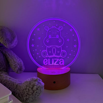 A personalized 3D optical illusion night light shaped like a hippo sitting in a circle, glowing with purple light and displaying the name 'eliza'. It has a round wooden base and is shown on a white table next to a gray plush toy.