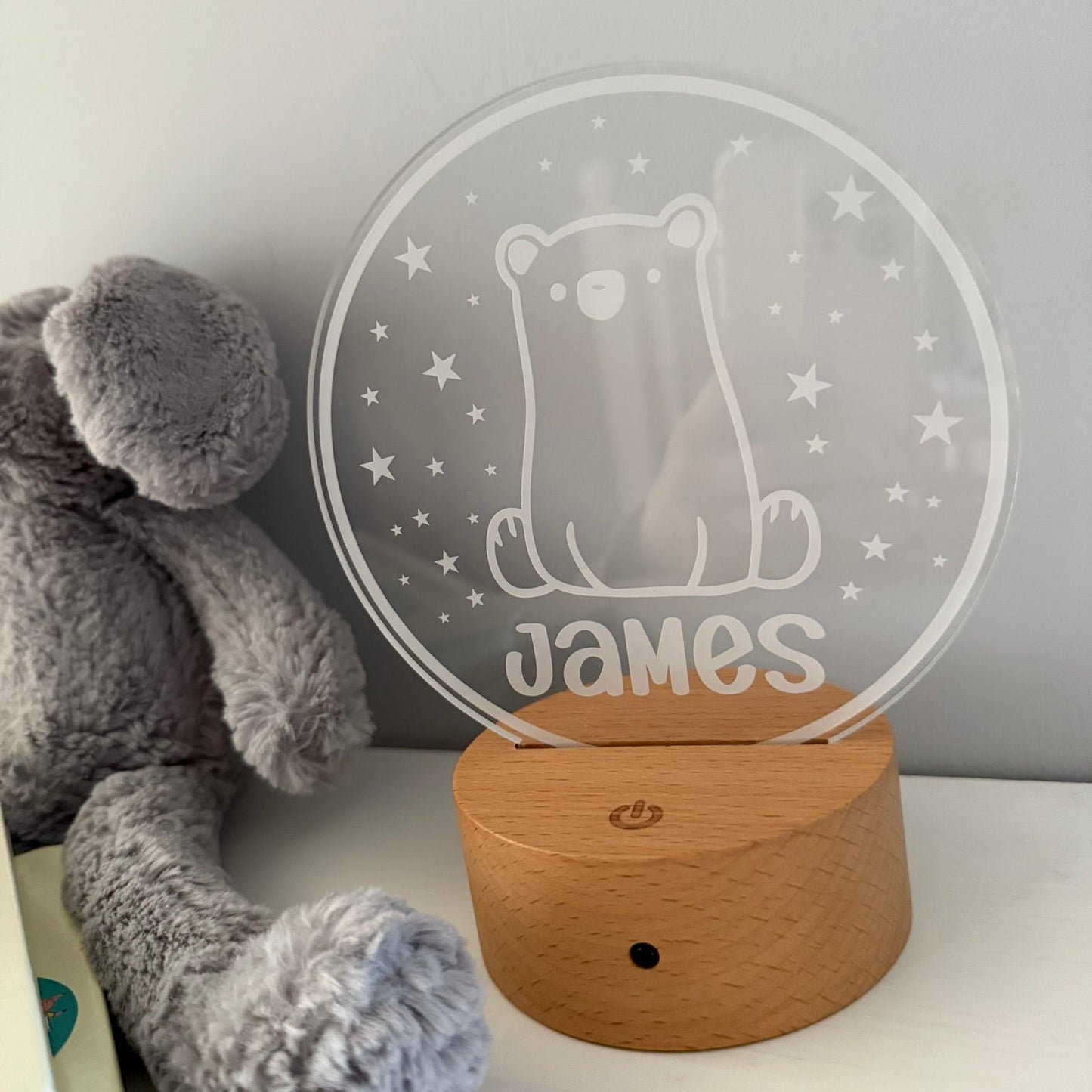 A personalized acrylic night light shaped like a bear sitting inside a circle with stars, engraved with the name 'James', resting on a round wooden base next to a gray plush toy.
