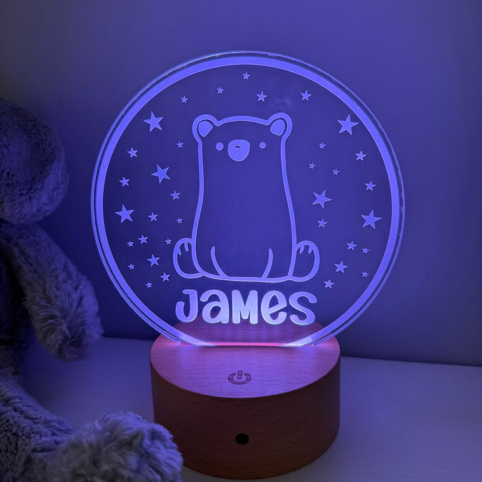 A personalized 3D optical illusion night light shaped like a bear sitting on a wooden base, glowing with a blue-purple light. The name 'James' is custom engraved at the bottom.