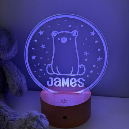 A personalized 3D optical illusion night light shaped like a bear sitting on a wooden base, glowing with a blue-purple light. The name 'James' is custom engraved at the bottom.
