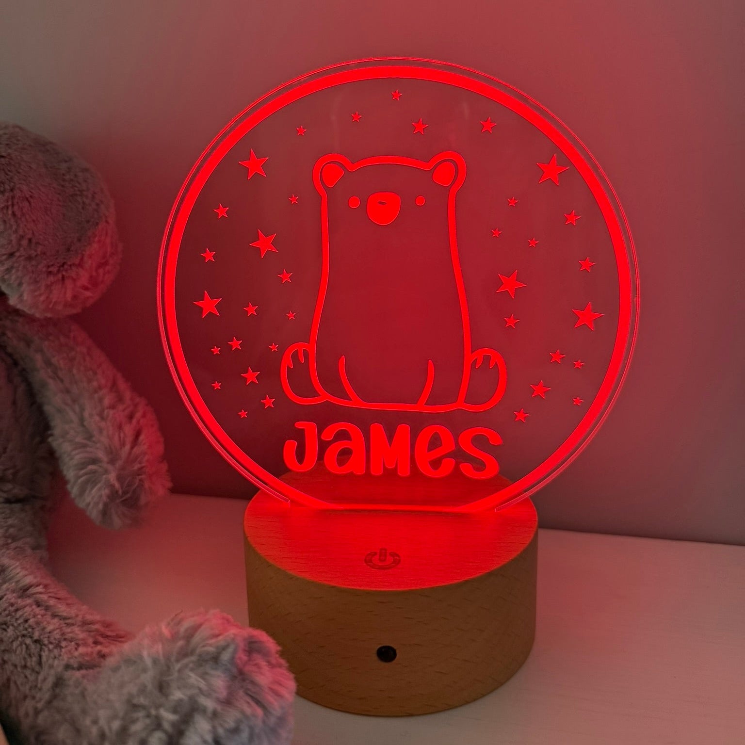 A personalized 3D optical illusion LED night light shaped like a bear sitting inside a circle, glowing red with the name 'James' cut out at the bottom. It sits on a wooden base next to a gray plush toy against a plain wall.