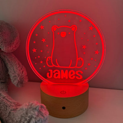 A personalized 3D optical illusion LED night light shaped like a bear sitting inside a circle, glowing red with the name 'James' cut out at the bottom. It sits on a wooden base next to a gray plush toy against a plain wall.