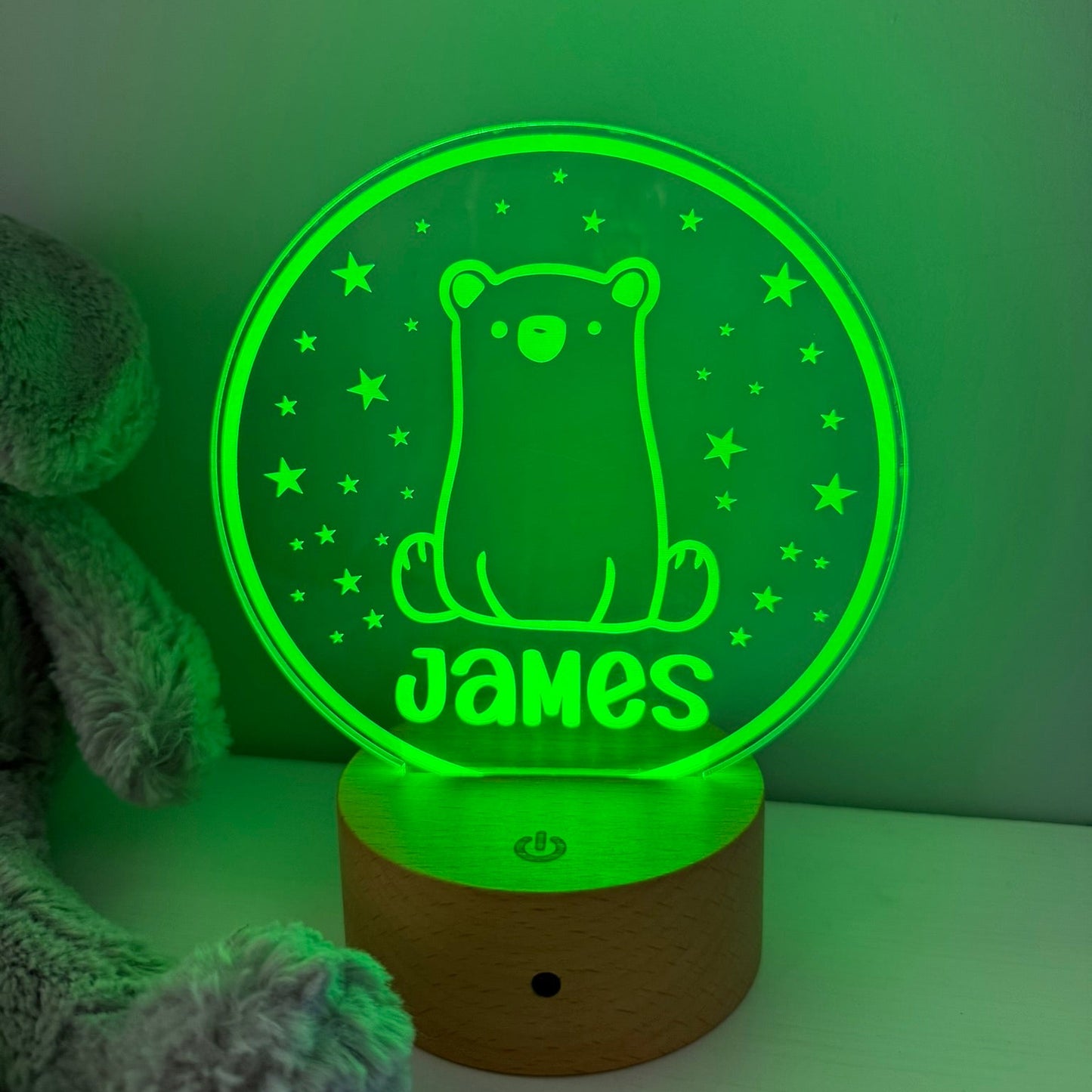 A personalized 3D optical illusion night light shaped like a round glowing disc on a wooden base, featuring a bear and the name 'James' illuminated in green. A plush grey stuffed animal is partially visible on the left against a green-lit background.