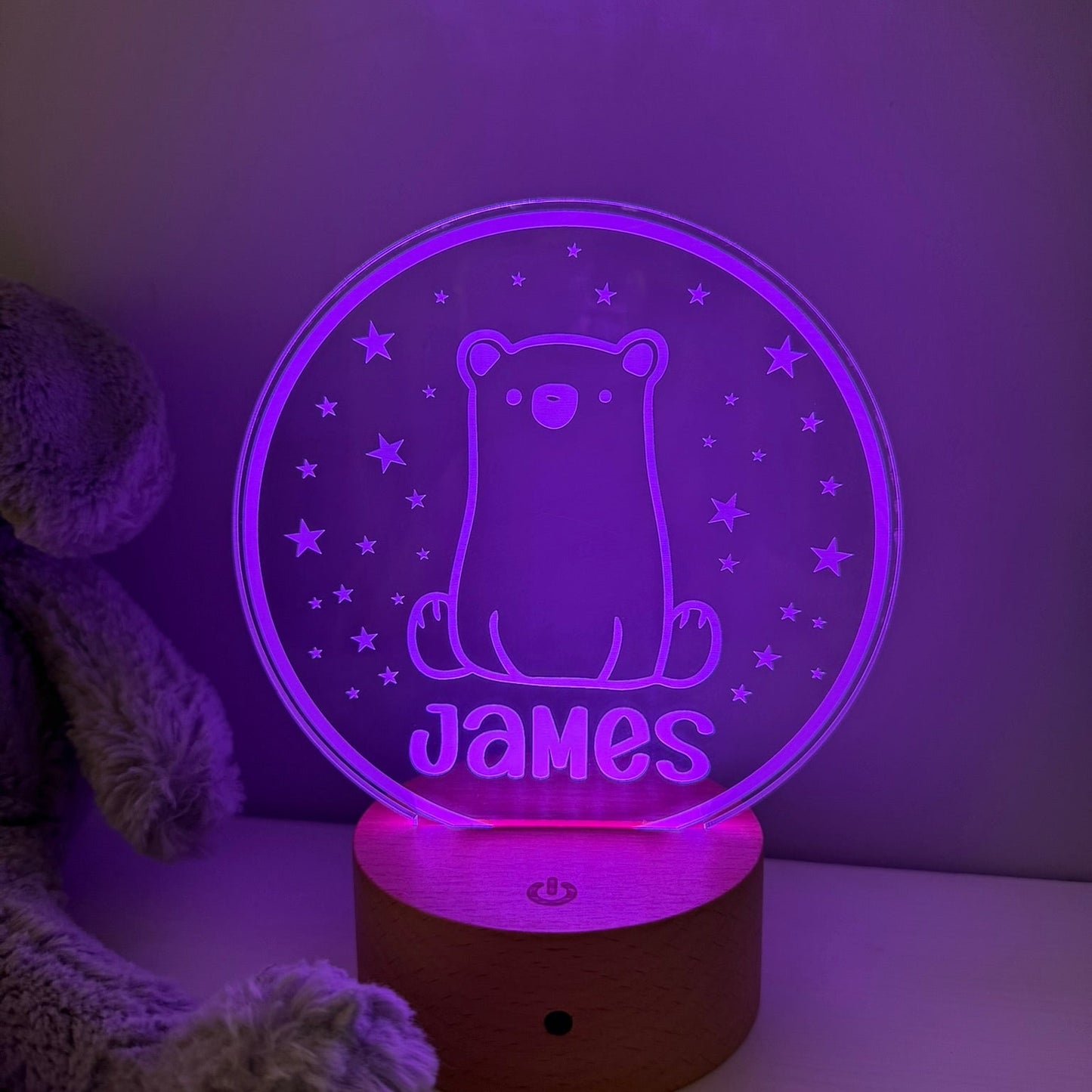 A personalized 3D optical illusion night light shaped like a circular clear plate with a glowing purple bear and the name 'James' on a wooden base, shown next to a plush toy in a dim room.