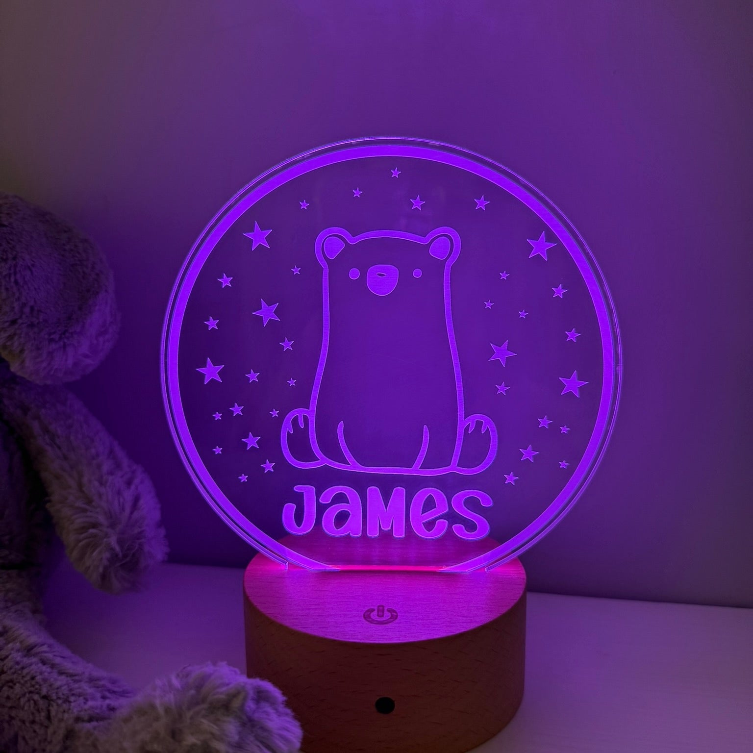 A personalized 3D optical illusion night light shaped like a circular clear plate with a glowing purple bear and the name 'James' on a wooden base, shown next to a plush toy in a dim room.