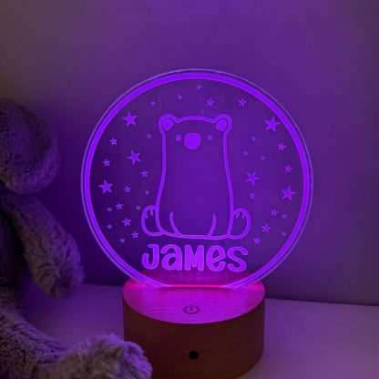 A personalized 3D optical illusion night light shaped like a circular clear plate with a glowing purple bear and the name 'James' on a wooden base, shown next to a plush toy in a dim room.