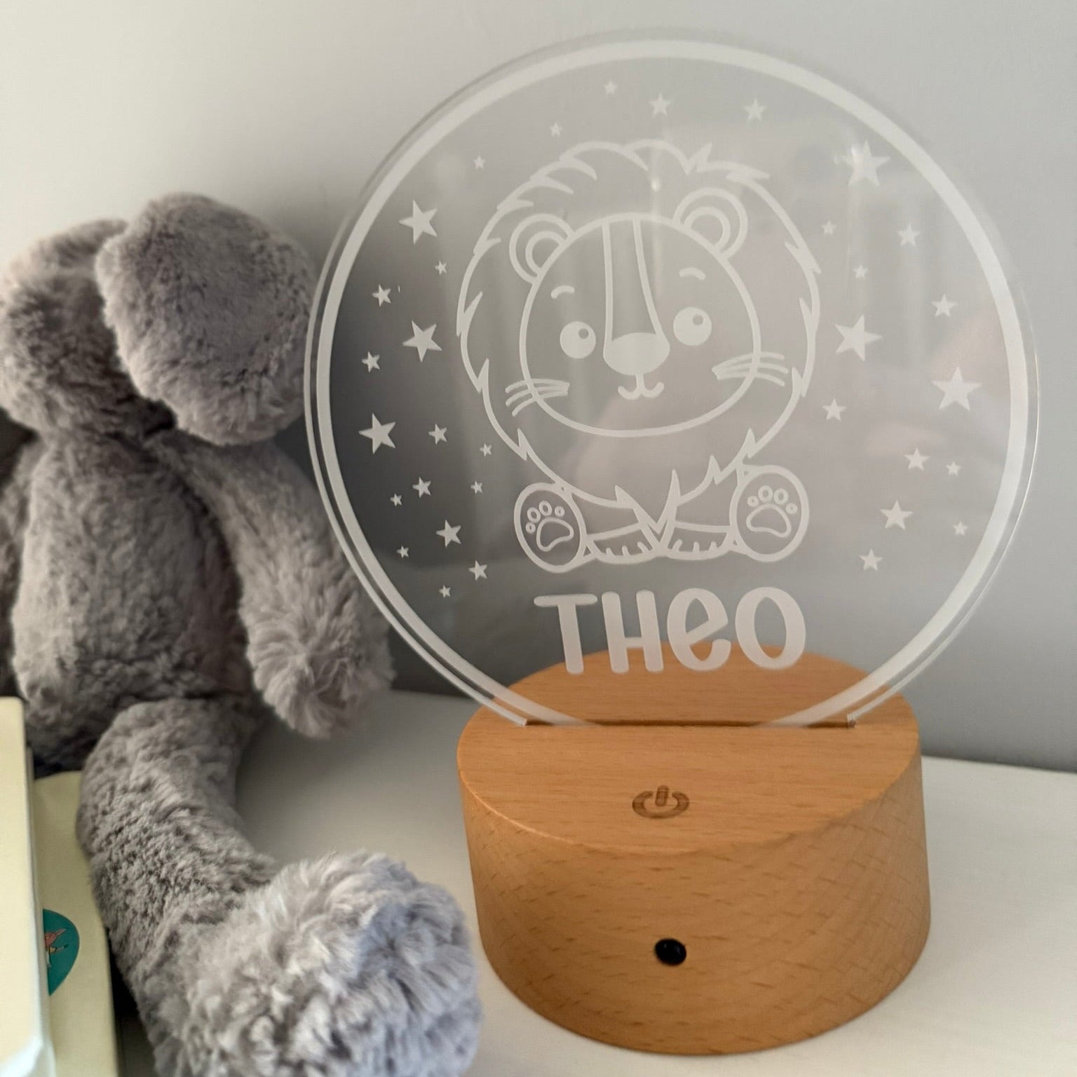A personalized LED night light with a wooden base and a clear acrylic disc featuring a lion illustration and the name 'THEO', shown next to a gray plush toy on a white surface against a light gray wall.