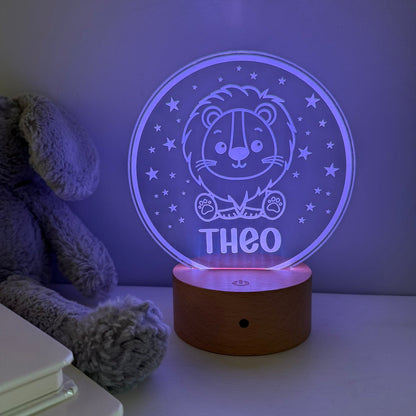 A personalized LED night light with a wooden base, displaying a lion illustration and the name 'THEO' in glowing purple light. The lamp is shown on a tabletop next to a gray plush toy and books, casting a soft purple glow on the wall.