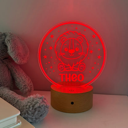 A personalized 3D acrylic LED night light shaped like a bear sitting in a circle, glowing red with the name 'THEO' and stars, resting on a wooden base next to a gray plush toy.