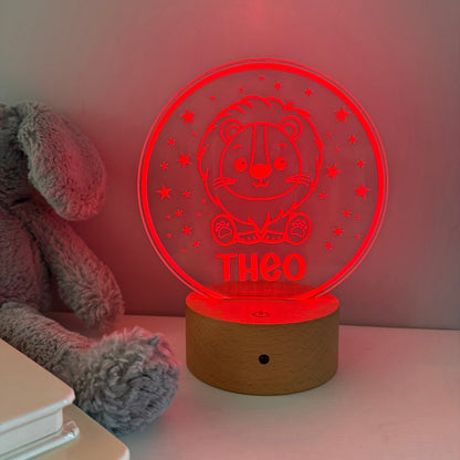 A personalized LED night light with a circular red-lit acrylic top featuring a teddy bear design and the name 'THEO', resting on a wooden base on a tabletop next to a gray plush toy.