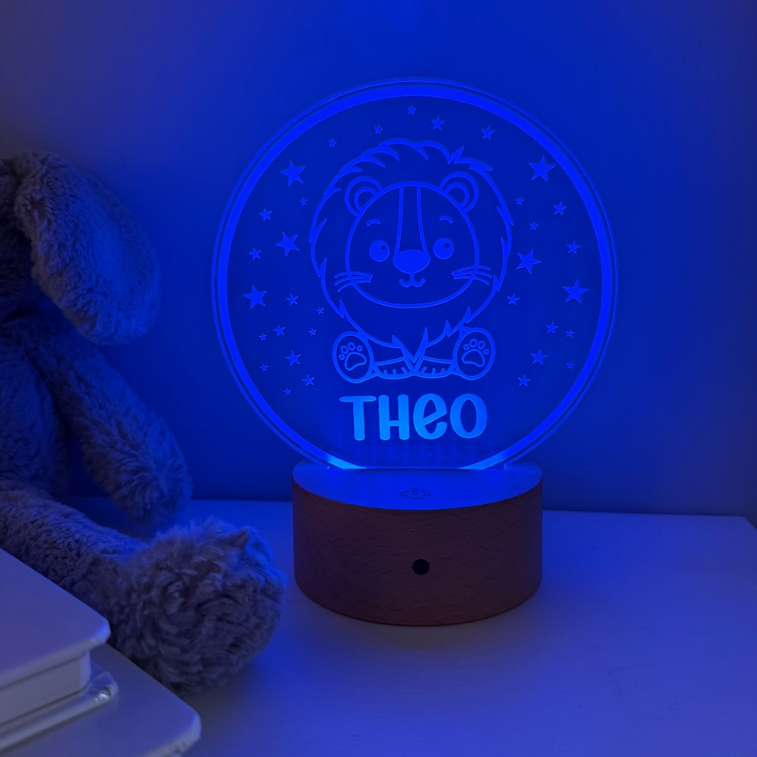 A personalized LED night light with a lion design and the name 'THEO' illuminated on a wooden base, shown on a tabletop with a blue glow. A plush toy is partially visible on the left.