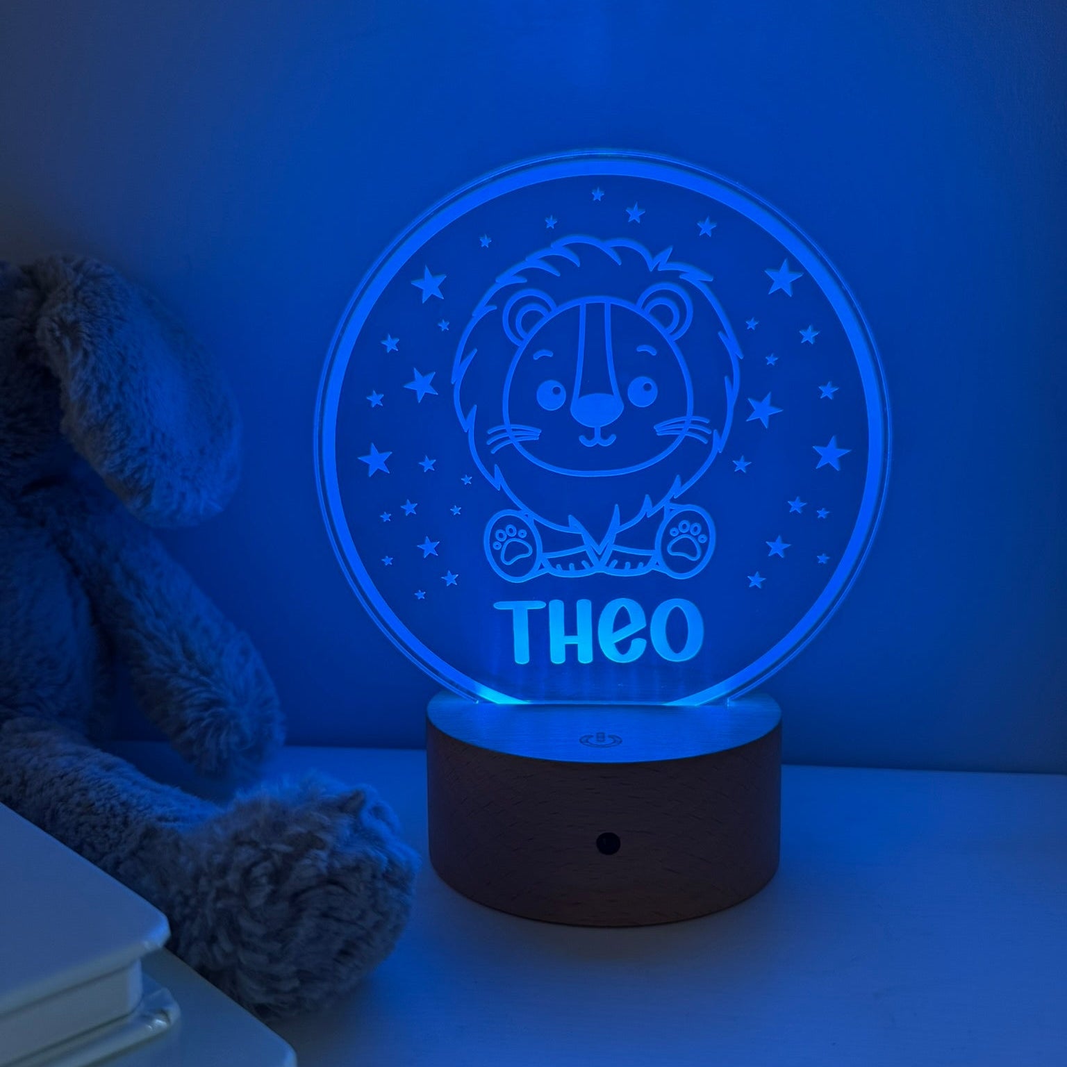 A personalized 3D optical illusion night light shaped like a circular wooden base with a glowing blue lion design and the name 'THEO' on the front, shown on a tabletop next to a teddy bear.